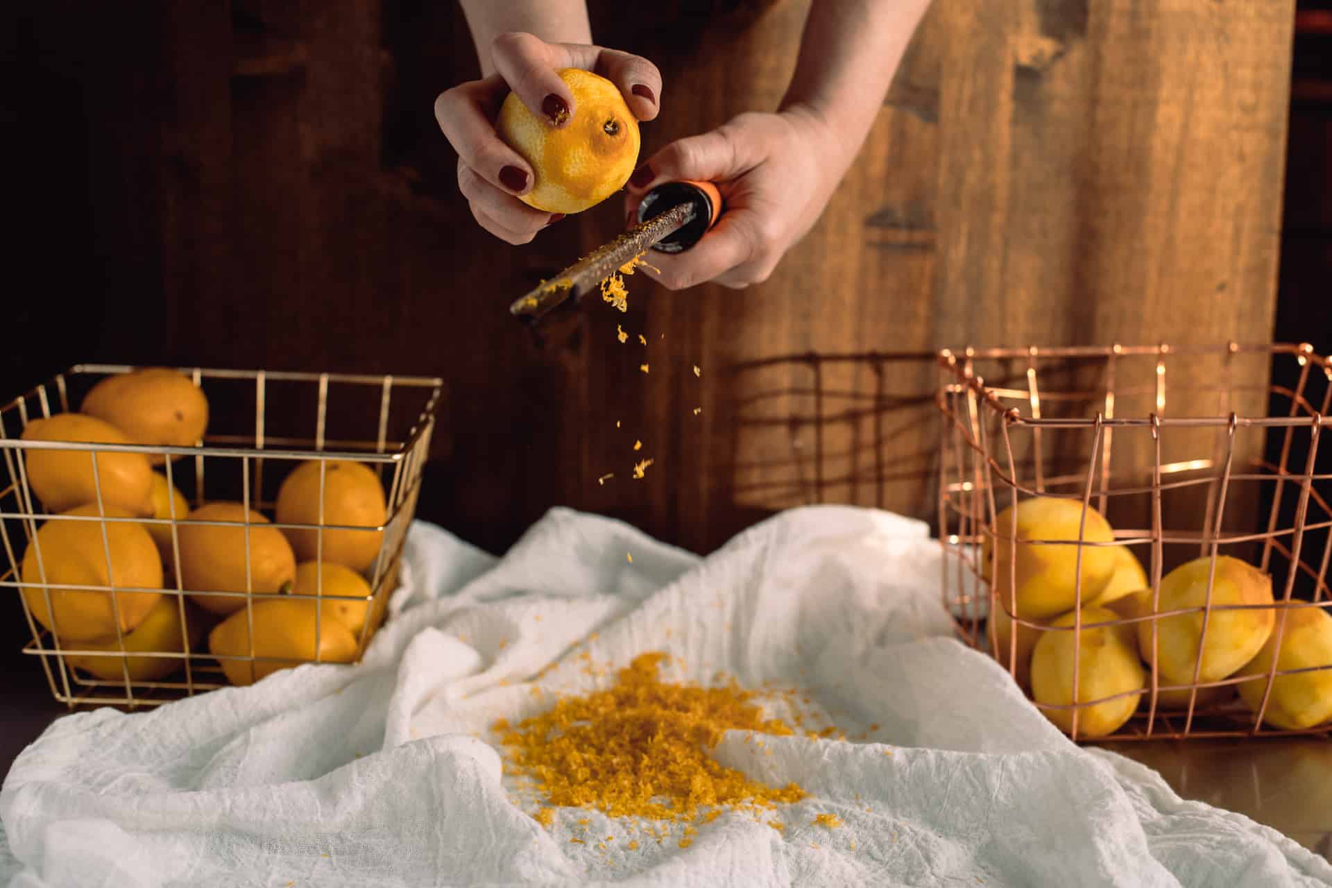 Hands zesting a fresh lemon over a microplane, copper baskets of lemons in a moody kitchen